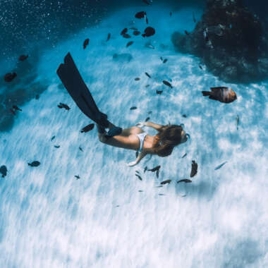 Free diver young woman with fins glides over sandy bottom with tropical fishes in transparent ocean